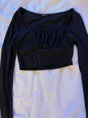 Svart croppad topp från SHEIN - Trendig svart croppad topp från SHEIN med långa ärmar och rynkad detalj framtill. Toppen har en fyrkantig urringning och är tillverkad i ett stretchigt material som sitter snyggt på kroppen. Perfekt för dig som gillar en stilren men ändå cool look.