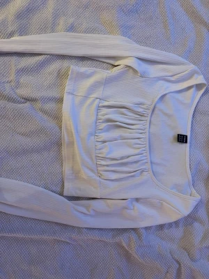 Vit ribbad croppad topp från SHEIN - Säljer en vit croppad topp från SHEIN med långa ärmar och ribbad struktur. Toppen har en snygg rynkning framtill och bred urringning som ger en trendig look. Perfekt att matcha med höga jeans eller kjol.