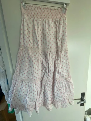 Vit långkjol med rosa blommor Gina Tricot - Söt långkjol från Gina Tricot i vitt med små rosa blommor. Kjolen har smock i midjan, volangdetaljer och är gjord i ett lätt och luftigt bomullstyg. Perfekt för en somrig och avslappnad stil.