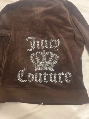 Brun velour hoodie Juicy Couture - Brun hoodie i mjuk velour från Juicy Couture med stora glittriga strasstenar som bildar loggan och en krona på ryggen. Hoodien har lång ärm och klassisk passform. Perfekt för dig som gillar en lyxig och avslappnad stil.