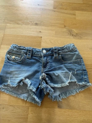 Blå low waist jeansshorts - Säljer ett par blå low waist korta jeansshorts. Shortsen har klassisk femficksdesign, bälteshällor och knappstängning framtill. Perfekta för varma dagar och en avslappnad stil. Ganska ”stora” i storlek.