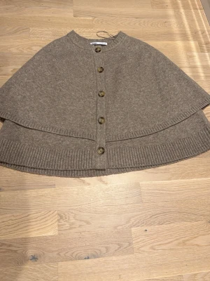 Beige stickad kofta från Zara - Säljer en beige stickad kofta från Zara med rund hals och stora knappar framtill. Koftan har ponchomodell och bred ribbad kant nedtill.