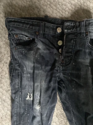 Mörkblå slitna skinny jeans från dsquard - Säljer ett par mörkblå skinny jeans med slitningar och hål på benen. Jeansen har flera knappar i gylfen och detaljer som ger en cool, använd look. Perfekta för dig som gillar streetstyle och en avslappnad vibe.