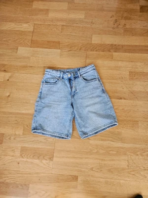Ljusblå denimshorts med normal passform - Säljer ett par klassiska ljusblå jeansshorts i denim med dragkedja framtill. Shortsen har en normal passform och midja, perfekta för en avslappnad stil under varma dagar.