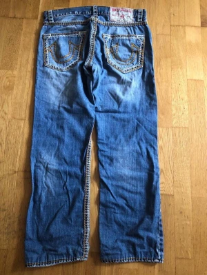 True Religion jeans med kontrastsömmar - True religion jeans med detaljerade sömmar i gult och vitt. Byxorna har en knappgylf och fem fickor, varav bakfickorna har True Religion-logotypens karaktäristiska design.  Modell: Ricky Super QT, storlek 33. Lite slitna i grenen (se foto).  Mått Midja: 44 cm Innerbenslängd: 73 cm Ytterbenslängd: 99 cm