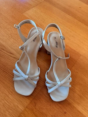 Beige klackskor med remmar. Aldrig använts - Snygga beige klackar  med smala remmar och justerbart spänne runt ankeln. Sandalerna har en låg klack och öppen tå, vilket ger en stilren och modern look. Tillverkade i ett slätt material som känns bekvämt på foten.