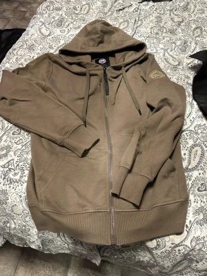 Brun hoodie med dragkedja Canada Goose - Säljer en brun hoodie från Canada Goose med dragkedja framtill och huva med dragsko. Märket syns tydligt på ärmen med ett broderat patch. Hoodien har ribbade muddar och är gjord i ett mjukt bomullsmaterial. Perfekt för en avslappnad och stilren look.