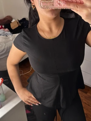 Svart peplum topp med kort ärm - Säljer en svart peplum topp med rund halsringning och korta ärmar. Toppen har en figurnära passform upptill och faller ut i en lätt volang nedtill. Perfekt att styla med jeans eller kjol för en trendig look.