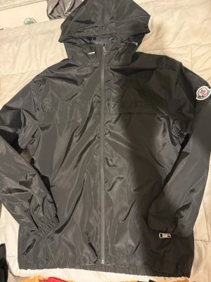 Svart vindjacka från Moncler - Snygg svart vindjacka från Moncler med huva och hel dragkedja framtill. Jackan har Moncler-logga på ärmen och elastiska muddar vid handleder och nederkant. Tillverkad i lätt, glansigt material som står emot vind. Perfekt för en stilren och sportig look.