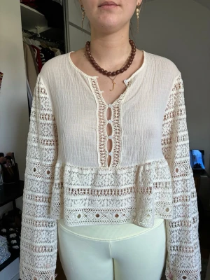 Vit boho-blus med spetsdetaljer - Supersöt vit blus i boho-stil med vida långärmar och vackra spetsdetaljer. Blusen har dekorativa knappar framtill och volang nedtill. Materialet är lätt och luftigt, perfekt för en trendig och avslappnad look.