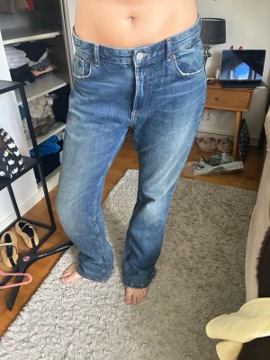 Blå raka jeans med hög midja - Säljer ett par klassiska blå jeans med rak passform och hög midja. Jeansen har fem fickor, ljus tvätt och subtila slitningar för en avslappnad look. Tillverkade i denim med snygga sömmar och knappgylf.