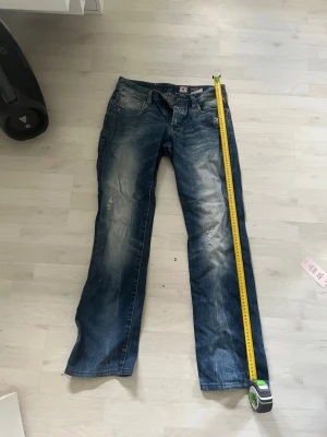 Blå slitna jeans med knappar - Säljer ett par blå jeans med slitningar och snyggt tvättad look. Jeansen har raka ben, klassiska fem fickor och knäppning med synliga metallknappar. Perfekta för en avslappnad och trendig stil.
