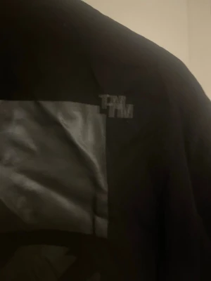 Svart t-shirt med glansigt tryck - Cool svart t-shirt med stort glansigt grafiskt tryck på ryggen och diskret logga vid axeln. T-shirten har klassisk passform och korta ärmar, perfekt för en streetwear-look.