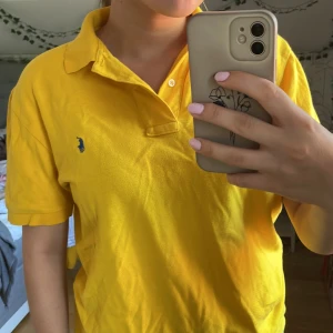 Polo by Ralph Lauren - Äkta Ralph Lauren, gul peké, kortärmad, knappar i kragen och lite uppskuret längst ner. Ingen skada förutom en liten missfärgning (se bild 3), och lätt missfärgning på insida krage (syns på bild 2)
