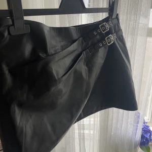 Kjol i skinnimitation  - Cool kjol i skinnimitation. Endast använd en gång därav är skicket som nytt. I och med att det finns spännen som detaljer går storleken att reglera en aning. 