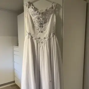 Säljer denna fina klänning som är precis som ny. Klänningen är vit med silverstenar. På kjolen är det blommor. Dragkedja i ryggen. Finns även en underkjol och handskar som ingår i priset. Finns inga defekter eller fläckar. Nypris 10 000. Strl M