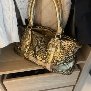 Guldig väska - Liten men ändå vääääldigt rymlig väska i guldigt ”krokodil mönstrat läder”. Väskan är använd, ooch det har lossnat lite typ på handtagen samt att innerfickans drag kedja är trasig. Men väskan ser ut som på bilderna! Hör av er vid förfrågan av fler bilder!