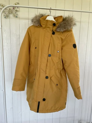 Vinterjacka  - En snygg lång, gul vinterjacka.  Bäde dragkedja och knappar som håller den tät.  4 st fickor 100 % polyester 