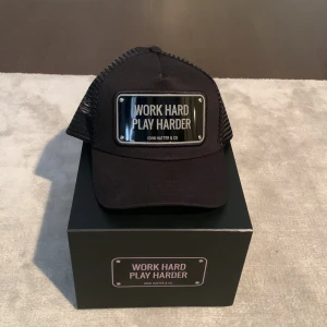 Svart John Hatter keps - Svart John Hatter keps med texten WORK HARD PLAY HARDER. Plåtbricka. Använd 1 gång.