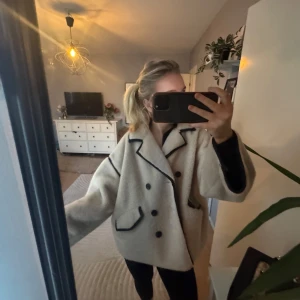 Beige teddyjacka med svarta detaljer - Säljer en mysig beige teddyjacka med svarta knappar och detaljer. Jackan har en oversized passform och långa ärmar, perfekt vår jacka. Den har en dubbelknäppt design och svarta kantlinjer som ger en snygg kontrast.