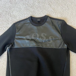 Svart tröja från Hugo Boss - Snygg svart tröja från Hugo Boss med en cool läderliknande panel över bröstet där loggan är diskret tryckt. Tröjan har långa ärmar och en rund halsringning. Perfekt för en stilren look.