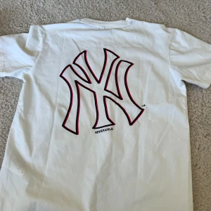Vit t-shirt med Yankees-logga - Snygg vit t-shirt från New Era med Yankees-logga i svart och rött på bröstet och en större version på ryggen. Perfekt för sportiga tillfällen eller som en stilren vardagslook.
