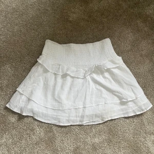 Vit kortkjol med volang - Söt vit kortkjol med volangdetaljer och elastisk midja. Perfekt för varma sommardagar. Kjolen har en luftig och bekväm passform.