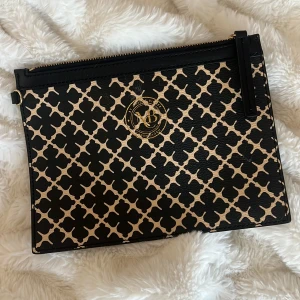 Malene Birger väska  - Snygg kuvertväska med ett unikt svart och beige mönster. Väskan har en praktisk dragkedja och ett elegant märke i guld på framsidan. Perfekt för att bära med sig det viktigaste med stil.