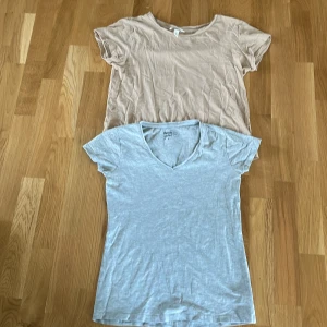 Två t-shirts i beige och grå - Säljer två enkla t-shirts i beige och grå. Den beige t-shirten har en rund hals och den gråa har en v-ringning. Perfekta för en avslappnad stil.
