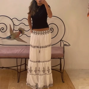 Vit långkjol med broderade detaljer - Elegant vit långkjol med vackra broderade mönster i svart. Kjolen har en knytning i midjan och flera dekorativa band som ger en bohemisk känsla. Perfekt för en stilfull och avslappnad look. Har bud på 260