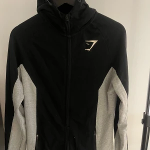 Svart och grå träningsjacka från Gymshark - Säljer en svart och grå träningsjacka från Gymshark. Jackan har en dragkedja framtill och är tillverkad i en blandning av polyester, bomull och elastan. Perfekt för träning och har en sportig look med långärmade ärmar.