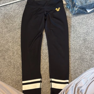 Svarta leggings från H&M Sport med Disney-motiv - Snygga svarta leggings från H&M Sport med ett litet guldigt Disney-motiv. De har vita ränder vid anklarna och är perfekta för träning eller en avslappnad dag. Tillverkade i ett stretchigt material för bästa komfort.