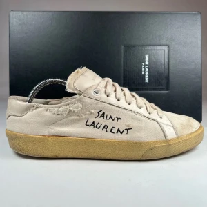 Vita sneakers från Saint Laurent - Snygga vita sneakers från Saint Laurent med en cool, sliten look. Skorna har snörning och en broderad logotyp på sidan. Perfekta för en avslappnad stil med en touch av lyx.
