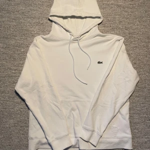Vit hoodie från Lacoste - Säljer en stilren vit hoodie från Lacoste med den klassiska krokodilloggan på bröstet. Hoodien har en justerbar dragsko i huvan och ribbade muddar vid ärmslut och nederkant. Perfekt för en avslappnad stil.
