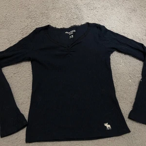 Svart ribbad tröja från Abercrombie Kids - Säljer en mörk grön ribbad tröja från Abercrombie Kids med v-ringning och långärmade ärmar. Tröjan har en liten broderad älgdetalj i vitt nedtill. Perfekt för en stilren look. Denna tröjan är stor i storleken och passar mig som är lång 13 år. 