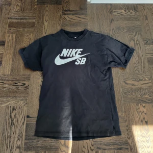 Svart t-shirt från Nike SB - Säljer en svart t-shirt från Nike SB med deras ikoniska logga i vitt på bröstet. T-shirten har en normal passform och korta ärmar. Perfekt för en avslappnad stil eller för skateparken.