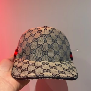 Beige och svart keps från Gucci - Snygg keps från Gucci i beige med svart GG-mönster. Kepsen har en klassisk design med justerbart band och röd-Mörkblå detaljer på sidan. Perfekt för att ge din outfit en lyxig touch. Pris kan diskuteras.