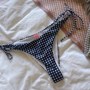 Blåvit rutig bikiniunderdel - Snygg bikiniunderdel i blåvitt rutigt mönster med knytband på sidorna. Perfekt för en dag på stranden eller vid poolen. Den har en klassisk och stilren design som passar till många olika överdelar.