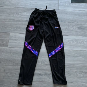 Svarta träningsbyxor från Nike med FCB-logga - Snygga svarta träningsbyxor från Nike med FCB-logga. Byxorna har en elastisk midja och färgglada detaljer i blått och rosa. Perfekta för fotbollsträningen eller en avslappnad dag.