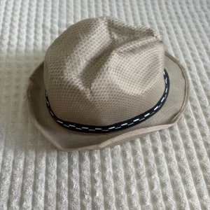 Beige hatt med svart band - Snygg beige hatt med ett svart band dekorerat med vita stygn. Perfekt för en stilren look. Hatten har en klassisk form och är tillverkad i ett lätt material.