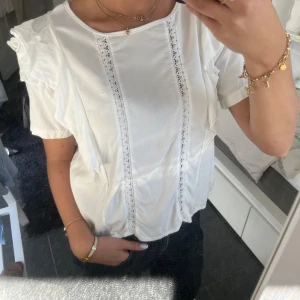 Vit blus med spetsdetaljer - Säljer en söt vit blus med spetsdetaljer framtill. Blusen har korta ärmar med volang och en rund halsringning. Perfekt för vår och sommar 💞