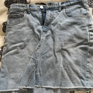Blå jeanskjol från Levi's - Snygg blå jeanskjol från Levi's med hög midja och klassisk femficksdesign. Kjolen har en rå kant nertill och en liten slits framtill för extra stil. Perfekt för en avslappnad look. Aldrig använd men finns ingen prislapp kvar