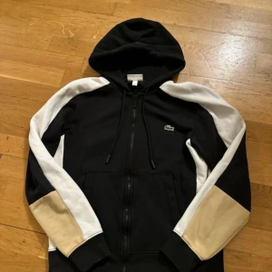 Svart hoodie från Lacoste - Säljer en snygg svart hoodie från Lacoste med dragkedja och klassisk passform. Tröjan har vita och beige detaljer på ärmarna och den ikoniska krokodilloggan på bröstet. Perfekt för en stilren look!