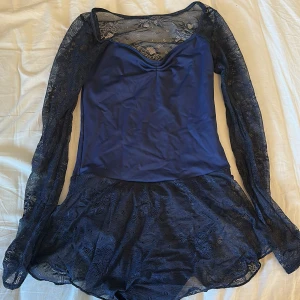 Marinblå spetsklänning - Elegant marinblå klänning med långa spetsärmar och en vacker spetsdetalj på ryggen. Klänningen har en figurnära passform och en kort kjol med volangkant. Perfekt för en speciell kväll!