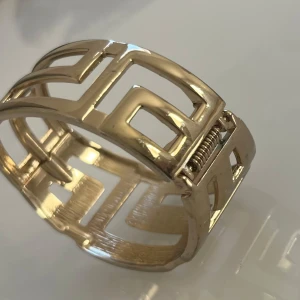 Guldfärgat armband med geometriskt mönster - Se gärna alla mina annonser, priserna kan diskuteras! Snyggt guldfärgat armband med ett stilrent geometriskt mönster. Armbandet har en öppen design som gör det enkelt att ta på och av. Perfekt för att ge en elegant touch till din outfit. Färg har släppts på vissa platser men inget man tänker på. Använd enstaka gång. 