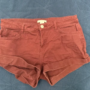 Röda lågmidjade shorts från H&M - Säljer ett par snygga kopparröda shorts från H&M. De har en klassisk design och är perfekta för sommardagar. Shortsen har en knapp och dragkedja framtill samt uppvikta benslut för en avslappnad look.