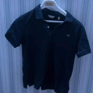 Svart kortärmad pikétröja från GANT - !behövs strykas!Säljer en stilren svart kortärmad pikétröja från GANT. Perfekt för en avslappnad men ändå snygg look. Tröjan har en klassisk krage och knappar framtill. Passar både till jeans och chinos.
