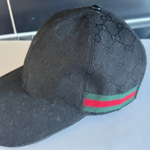 Svart keps från Gucci - Snygg svart keps från Gucci med diskret mönster och klassiska gröna och röda ränder på sidan. Kepsen har en justerbar rem baktill för perfekt passform. Den är använd men har mycket kvar att ge. 