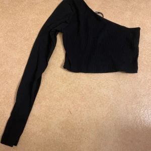 Svart oneshoulder topp - Snygg svart oneshoulder topp med ribbad textur. Perfekt för en trendig look. Den har en lång ärm och är croppad för en modern stil.
