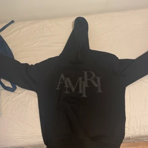 Svart hoodie från Amiri - Säljer en stilren svart hoodie från Amiri med stor logga på framsidan. Perfekt för en casual look med en skön passform och huva. Ett måste för garderoben!